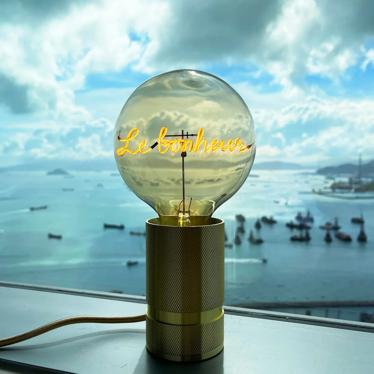 Ampoule Message in the Bulb verre ambré message Le Bonheur sur socle doré devant fenêtre avec vue sur la mer et les bateaux