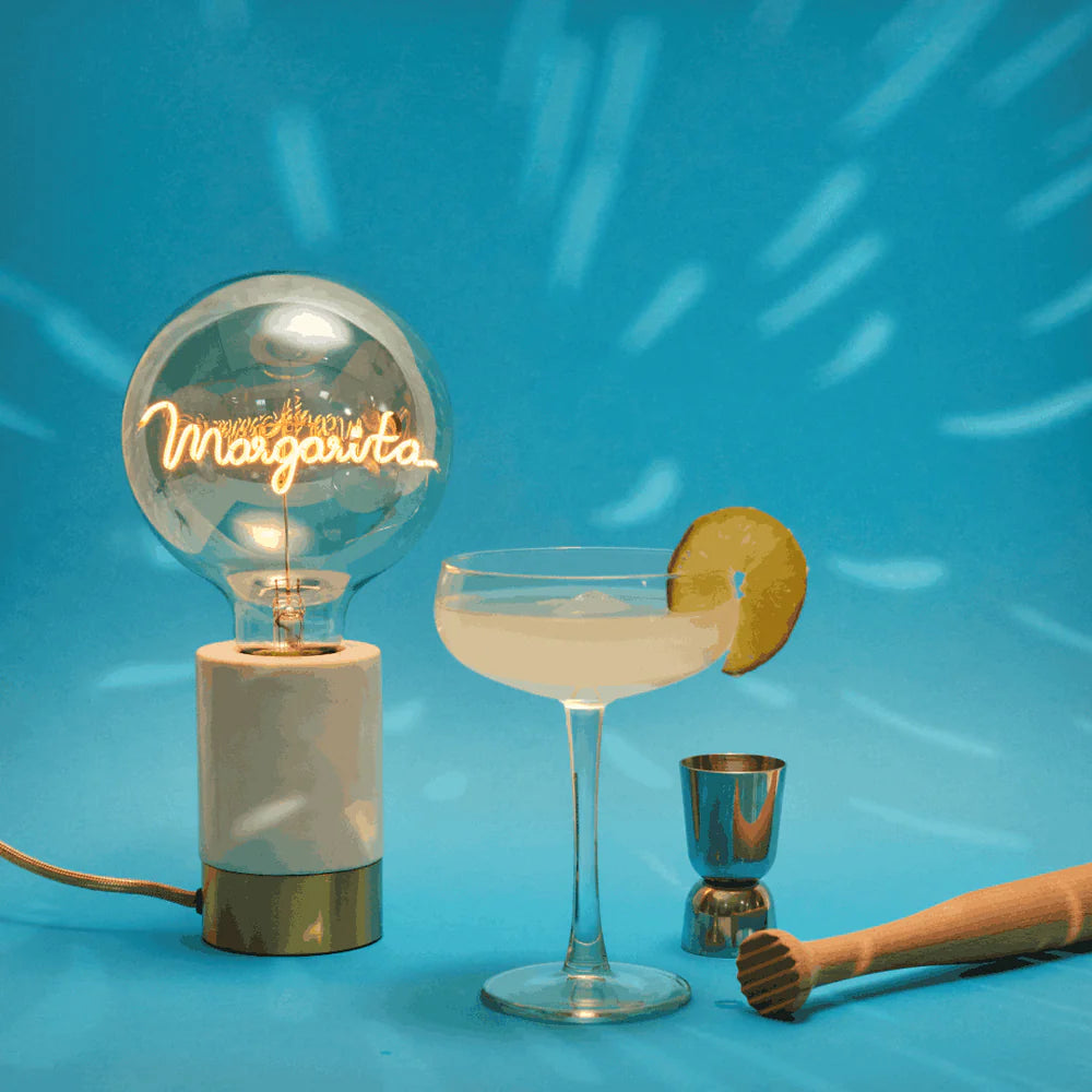 Ampoule Message in the bulb avec le mot Margarita écrit dans un style manuscrit qui s'illumine présentée avec un verre de Margarita et des accessoires de cocktail