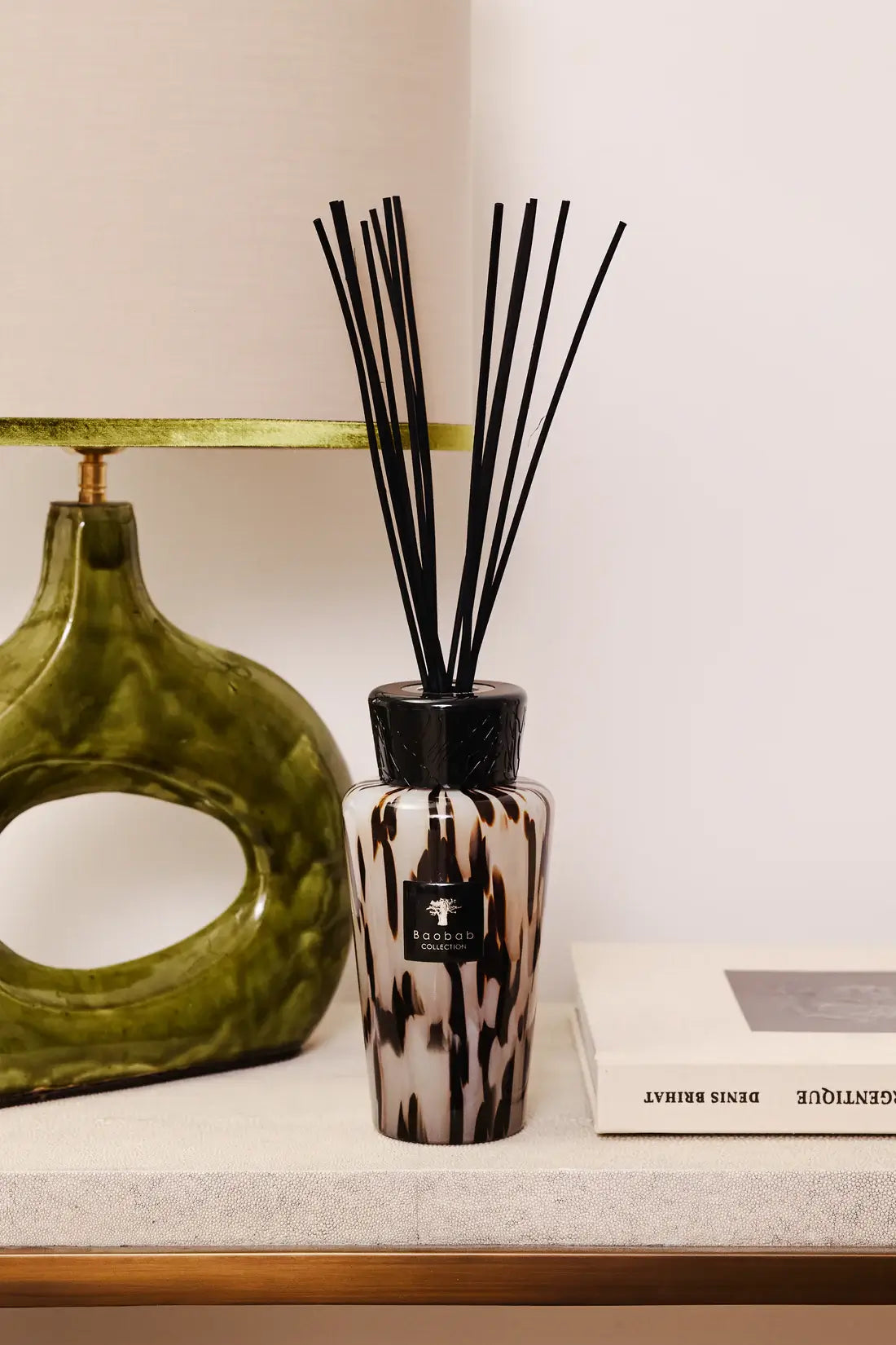 Baobab diffuseur Black Pearls 500ml sur une étagère à côté d'une lampe au socle vert et d'un livre de Denis Brihat