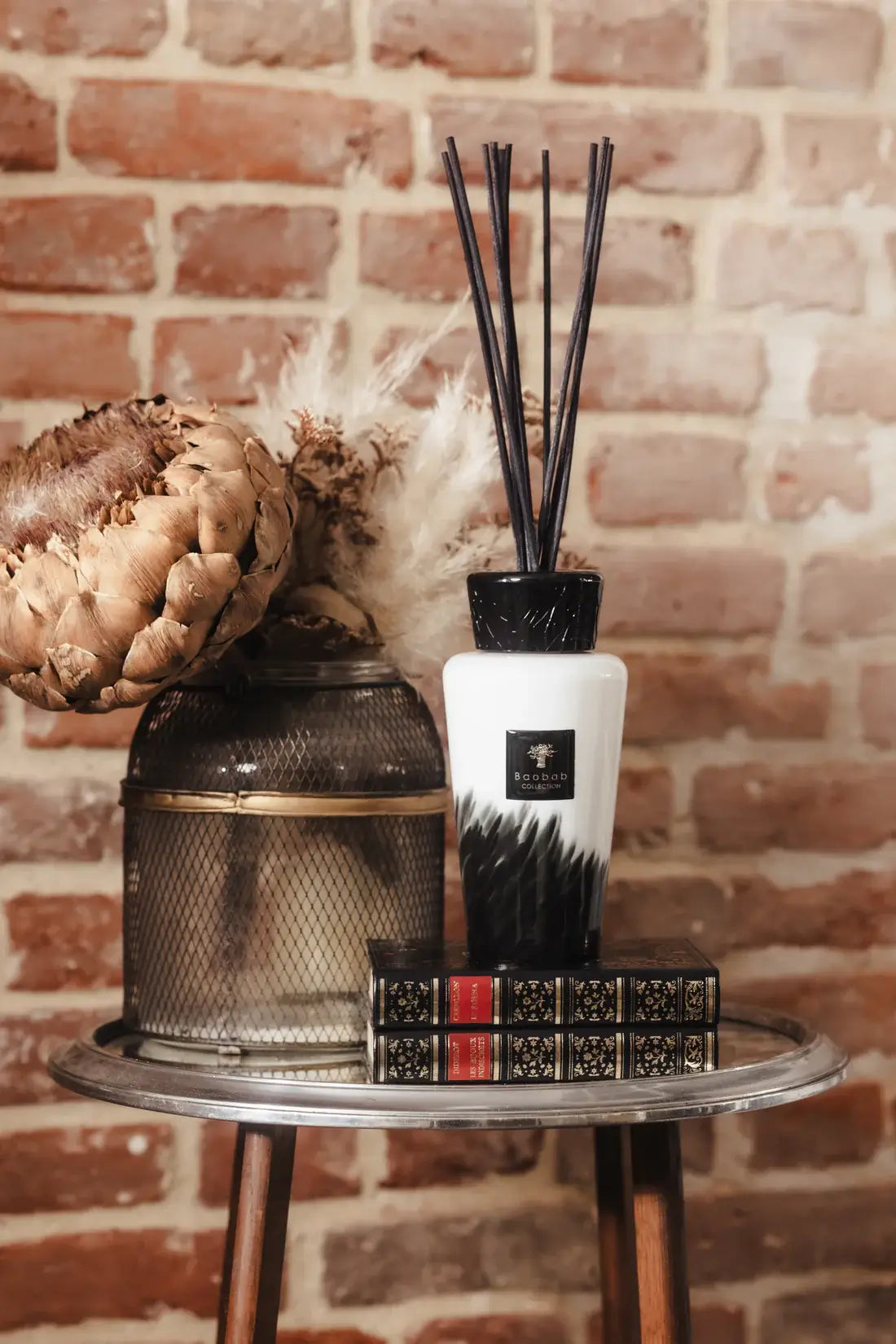 Baobab diffuseur Feathers 500ml dans un salon sur une table en verre avec un fond de mur de briques. Les motifs de la collection Feathers rappellent des plumes noires sur un fond blanc