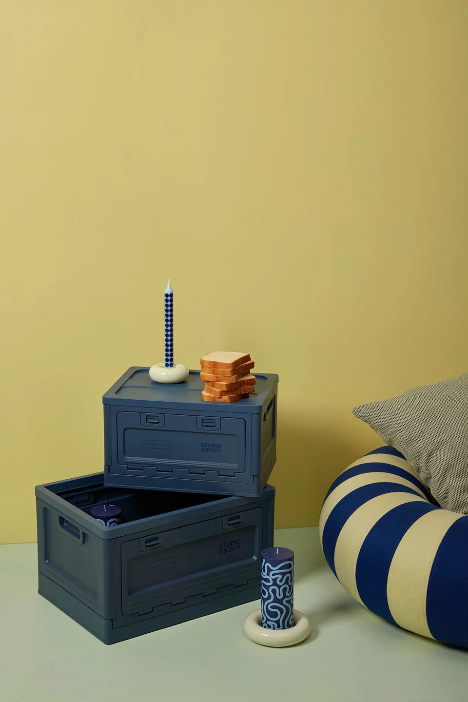 Les boites Studio About bleu foncé de taille medium et large empilés l'un sur l'autre. Avec la bougie pilier bleu foncé et le pouf bleu et jaune.