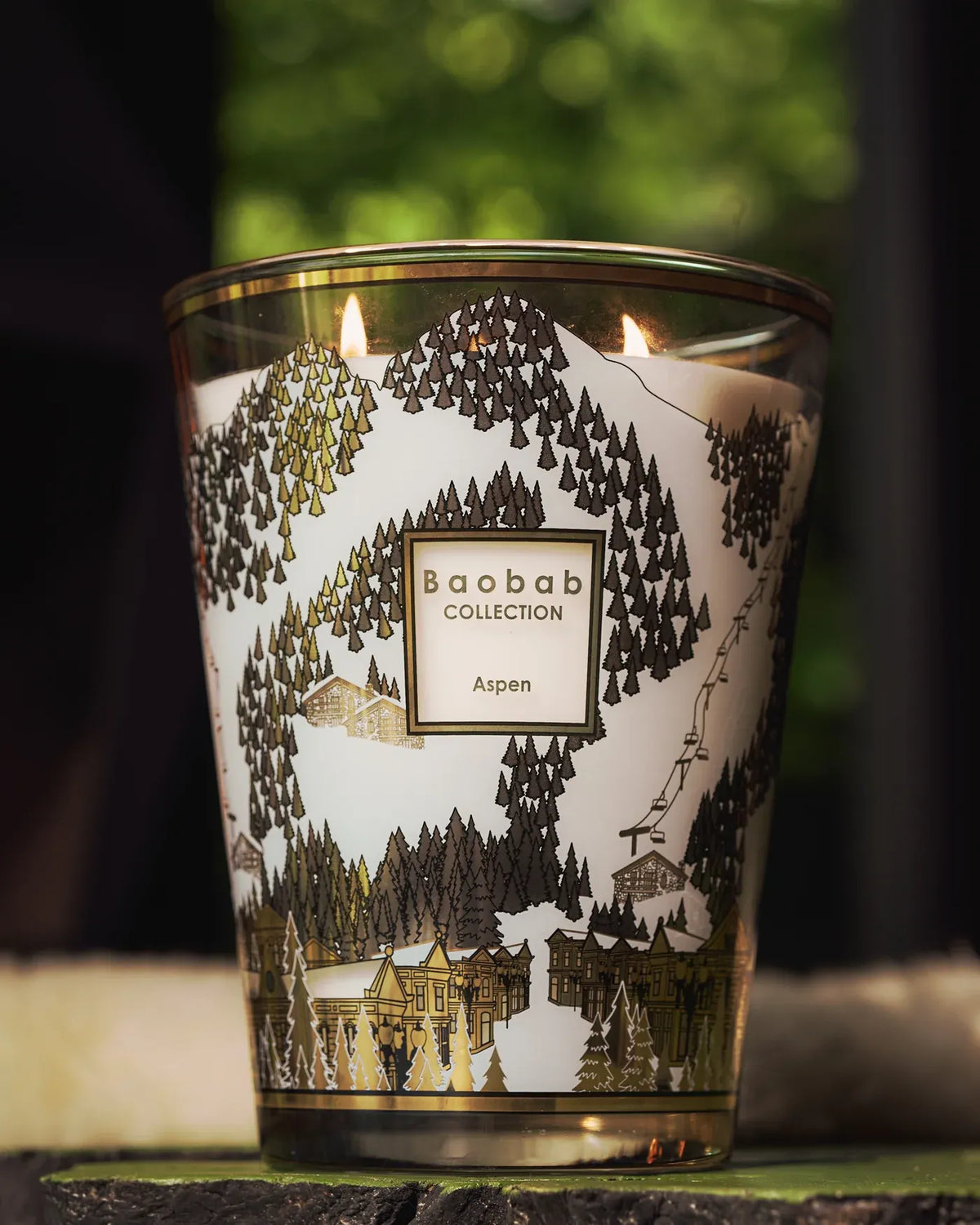 Bougie parfumée Aspen Baobab Collection allumée, décor de forêt de sapins et village alpin