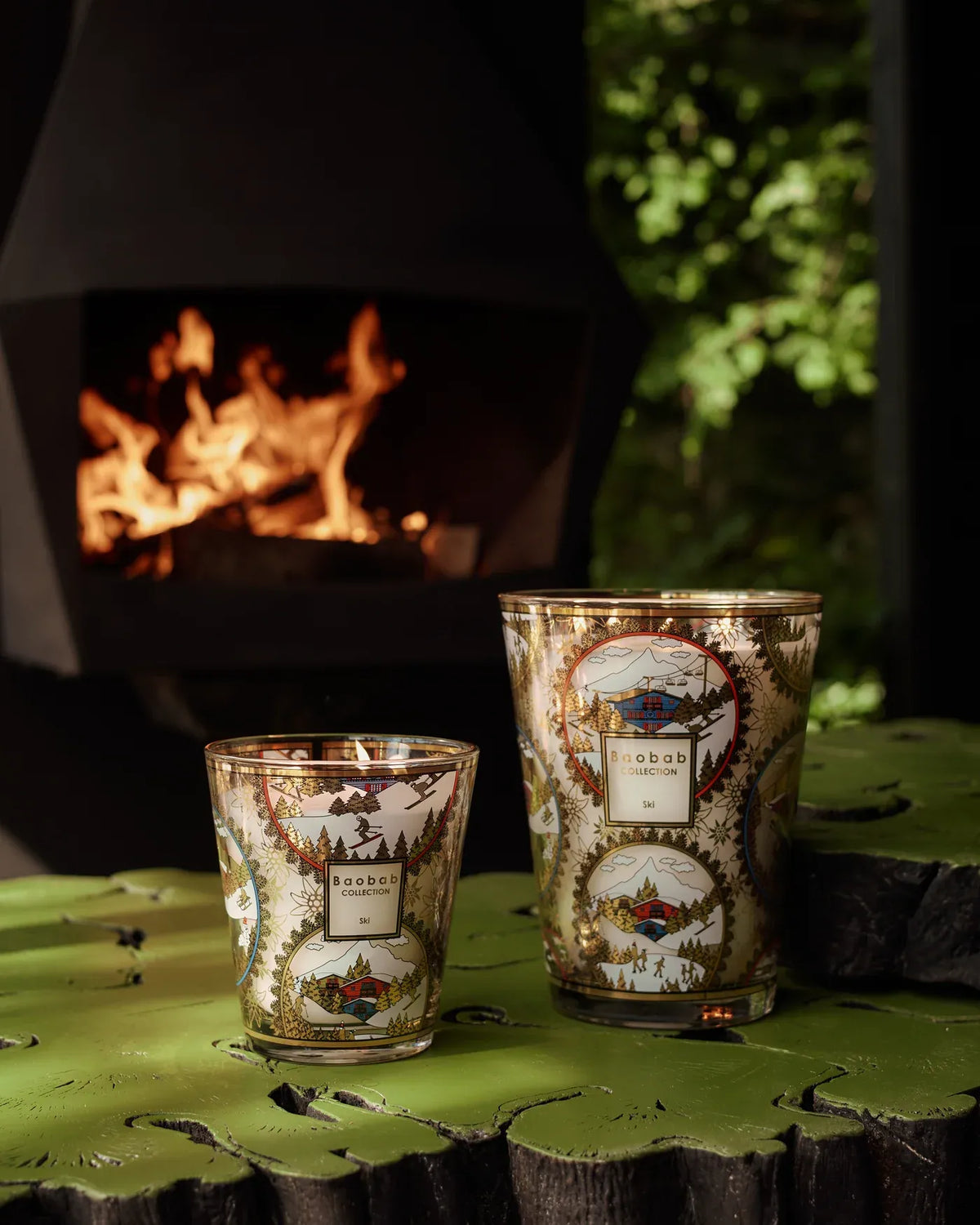 Bougie parfumée Ski Baobab Collection près d’un feu, décor illustré de chalets et de pistes enneigées