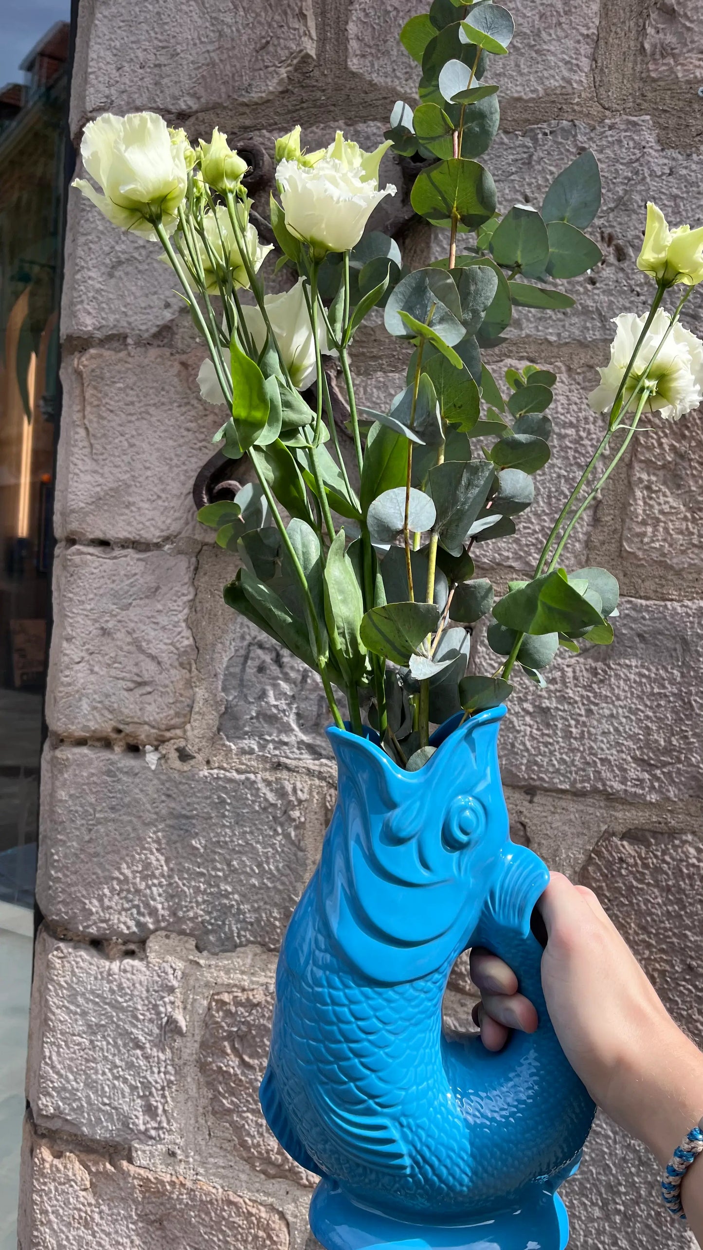 Vase Glouglou turquoise avec des fleurs, devant un mur en pierre 