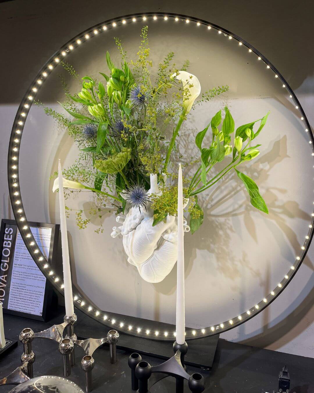 Vase représentant un cœur de chez Love In Bloom, fleuri avec des fleurs fraiches, entouré par un cercle de LED et derrière deux cierges contenant chacun une bougie.