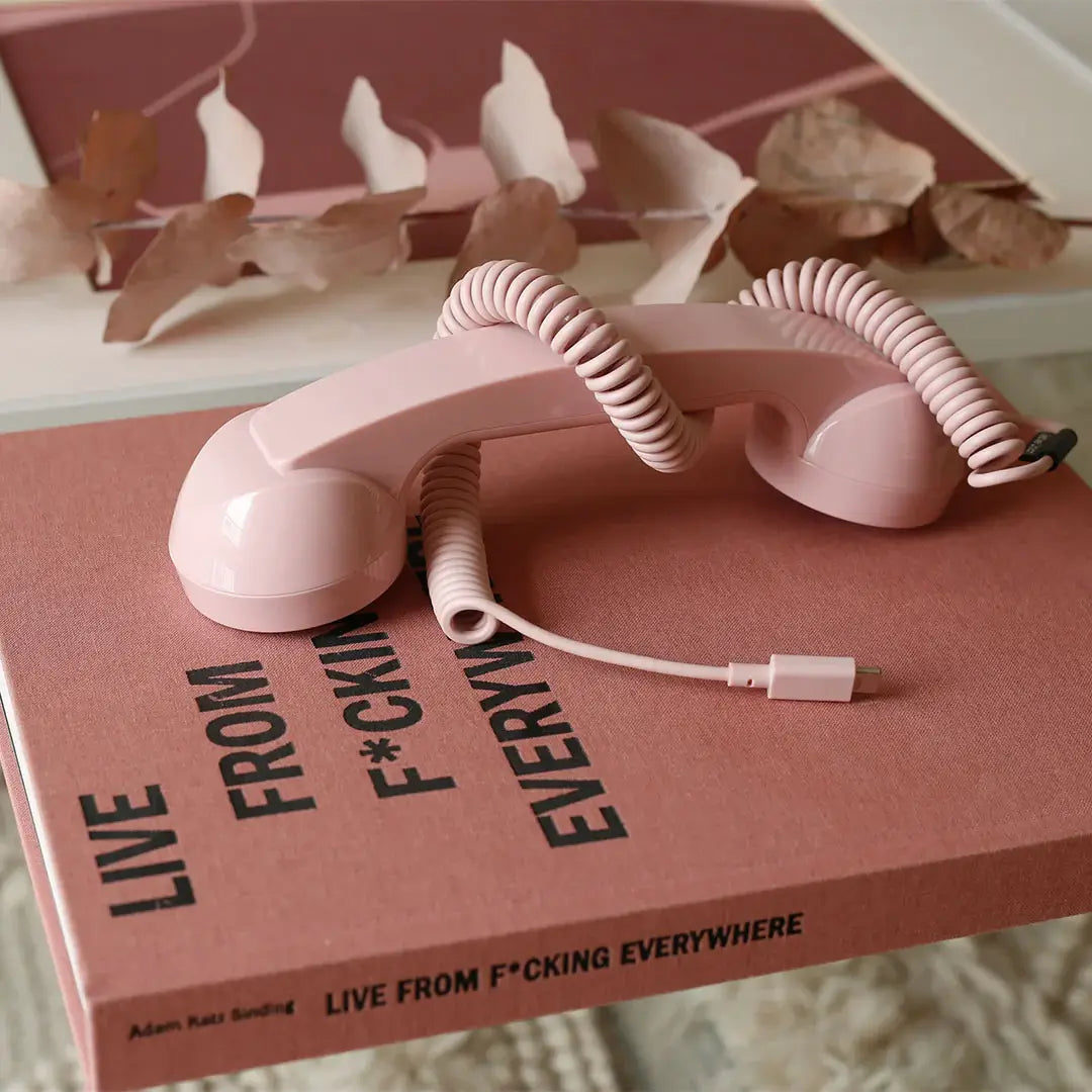 Combiné de téléphone Pop Phone Rose Bonbon de chez Native Union, posé sur un livre rose nommé "Live From F*cking Everywhere"