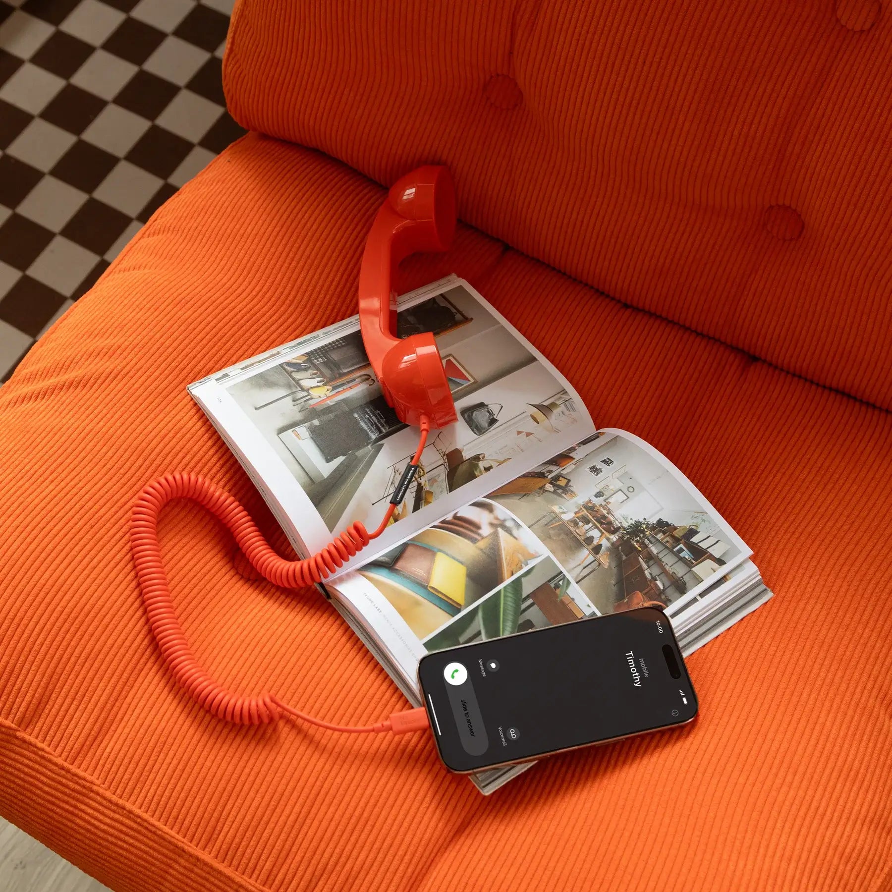 Combiné de téléphone Rouge Alarme Pop Phone de chez Native Union. Branché à un smartphone avec un câble USB-C. Le tout est placé sur un livre de décoration, sur un canapé rouge.